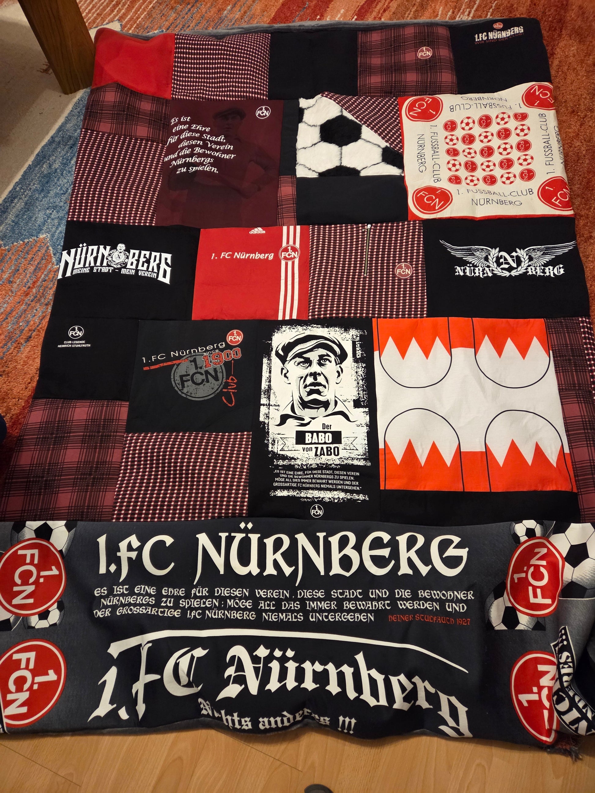 Eine Patchworkdecke mit Fußball-Motiven, Logos vom 1. FC Nürnberg und Frankenfahne liegt auf dem Boden ausgebreitet. Die Farben sind schwarz, weiß, rot, bordeaux.