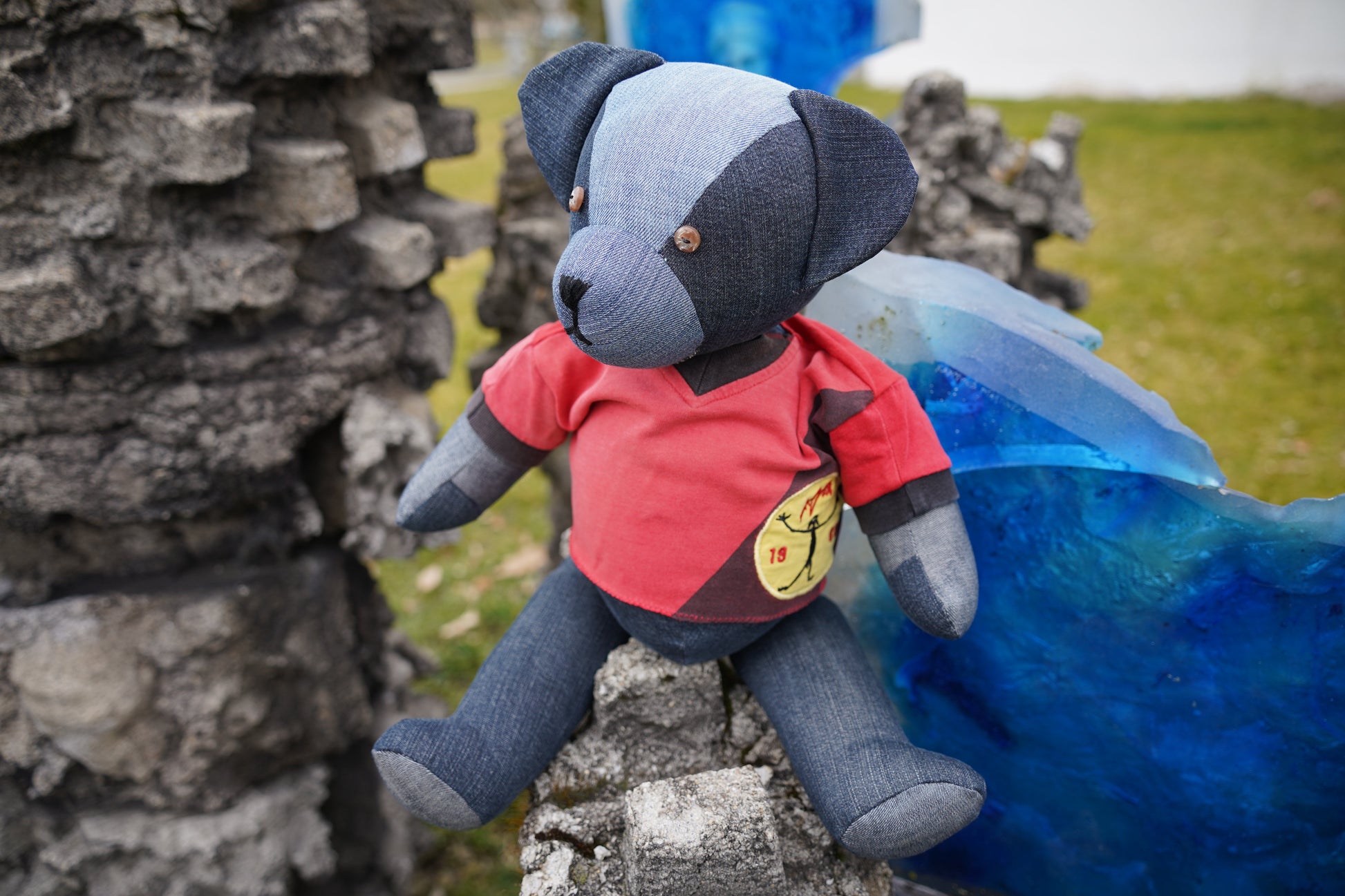 Ein blauer Teddybär aus Jeansstoff, der ein rotes Shirt trägt. Der Bär sitzt auf einer Steinmauer. Er hat braune Knopfaugen und eine schwarze Nase.