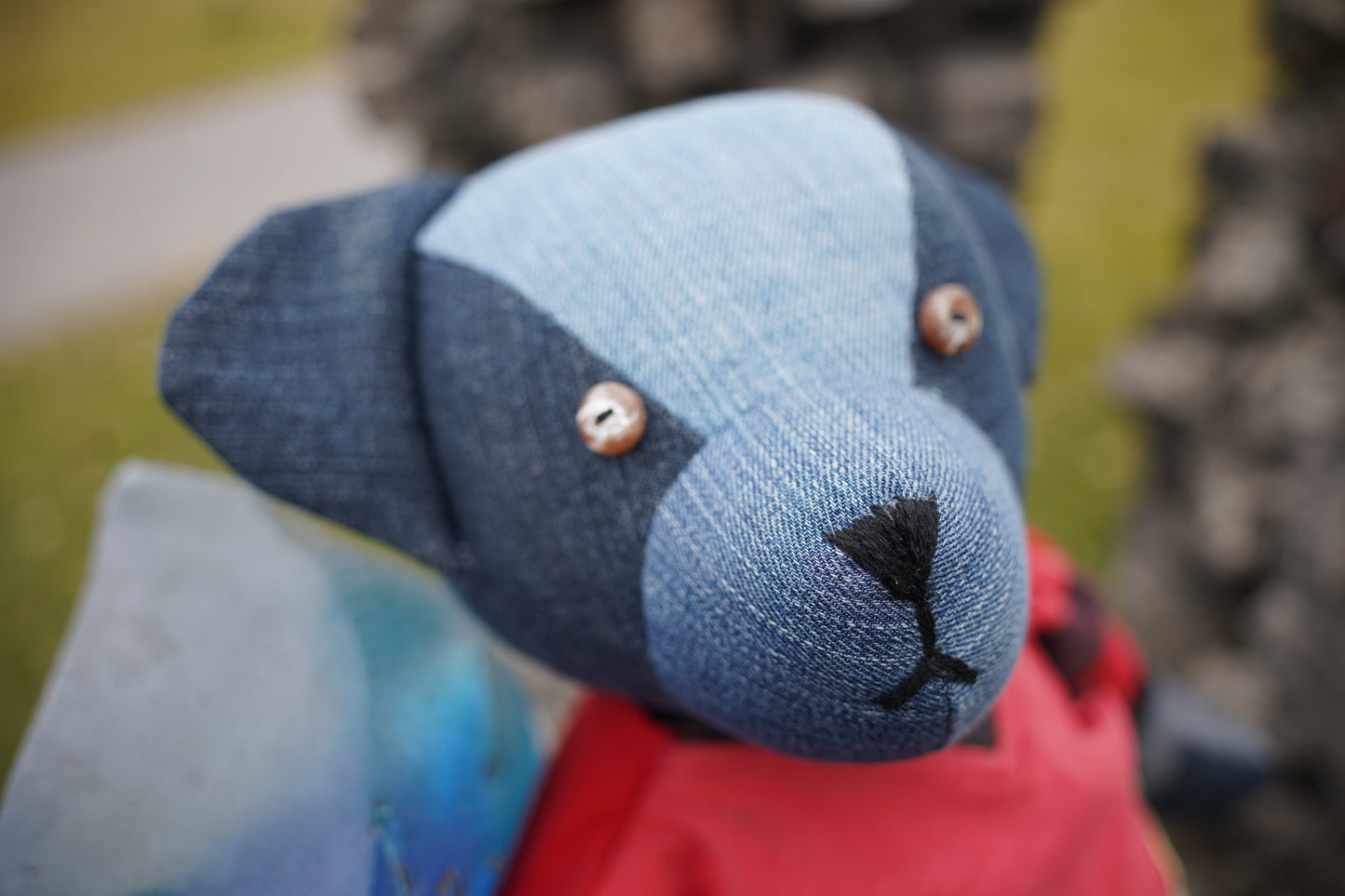 Der Kopf von einem blauen Teddybär aus Jeansstoff mit Knopfaugen und einer schwarzen gestickten Nase. Der Bär trägt ein rotes Hemd.