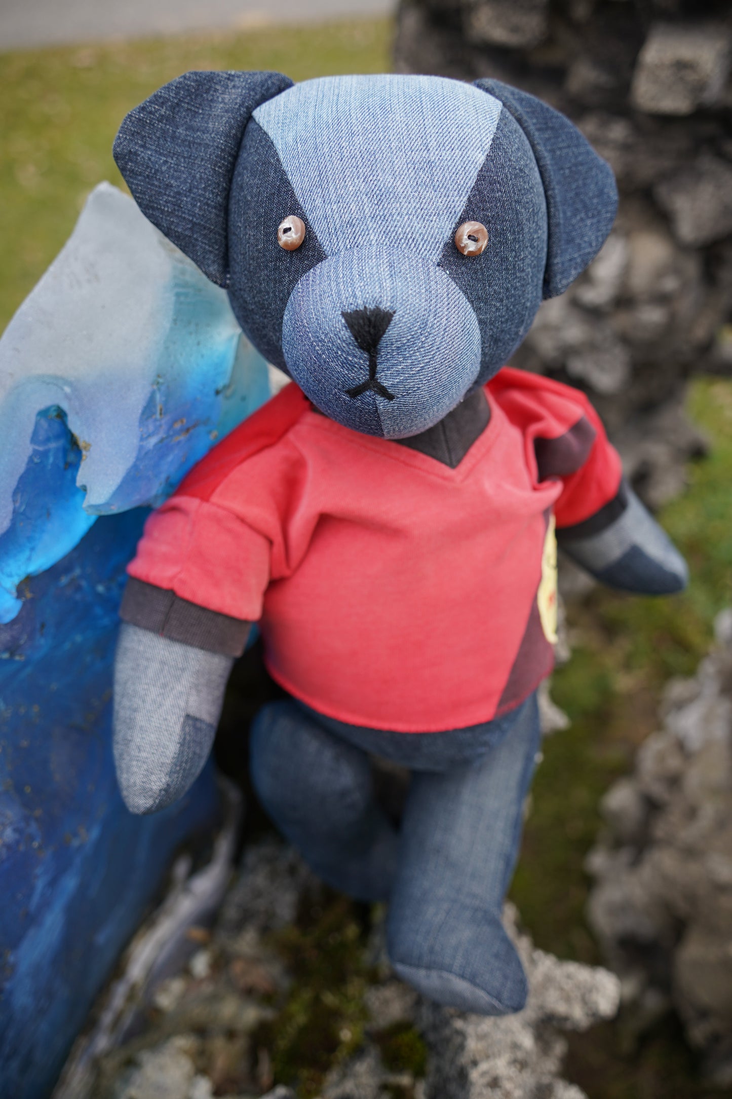 Ein blauer Teddybär aus Jeansstoff, der ein rotes Shirt trägt. Der Bär steht und hat braune Knopfaugen und eine schwarze Nase.