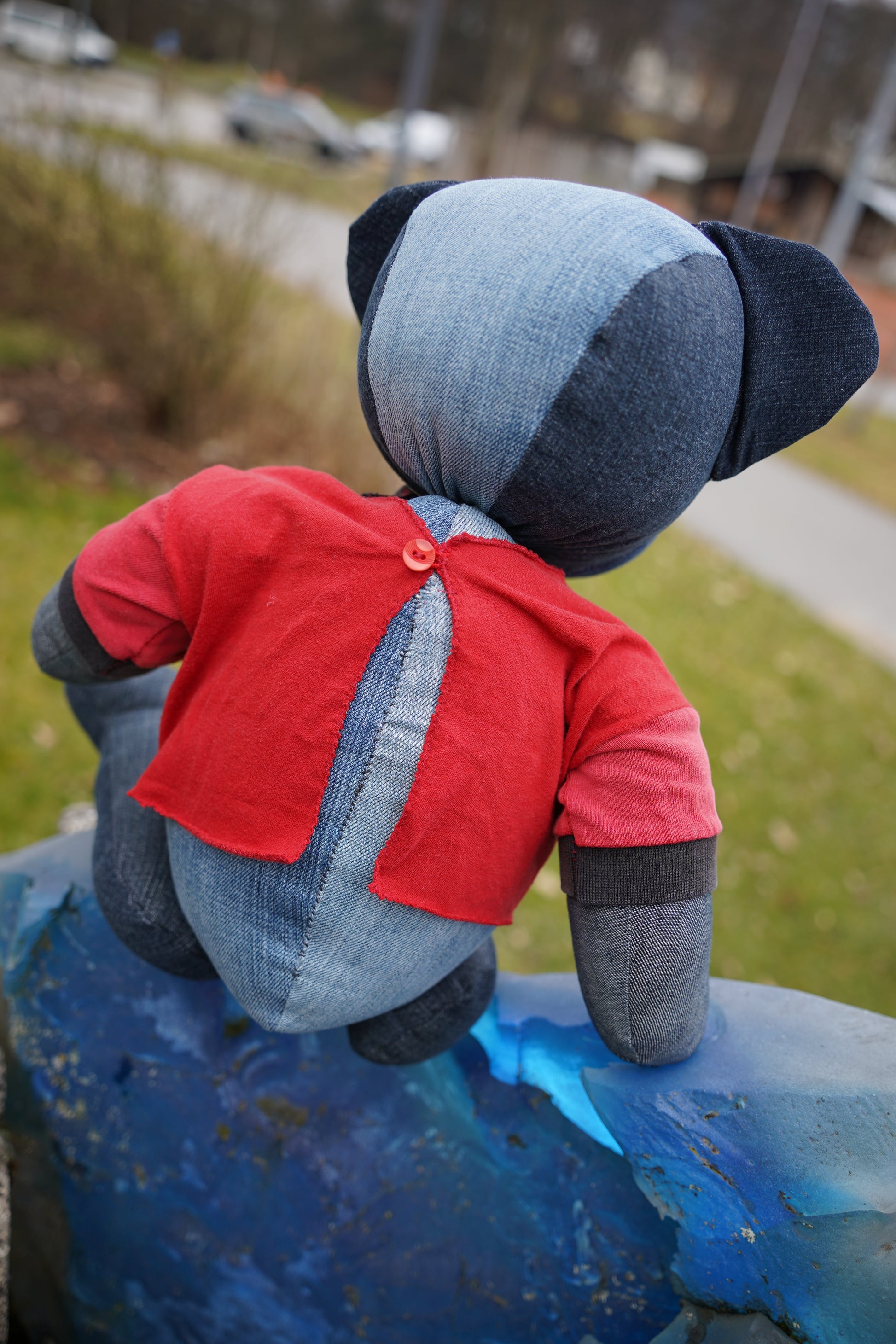 Ein blauer Teddybär von hinten aus Jeansstoff, der ein rotes Shirt trägt. Der Bär sitzt.