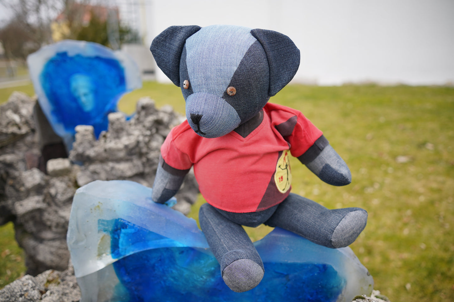 Ein blauer Teddybär aus Jeansstoff, der ein rotes Shirt trägt. Der Bär sitzt auf einem blauen Stein. Er hat braune Knopfaugen und eine schwarze Nase.