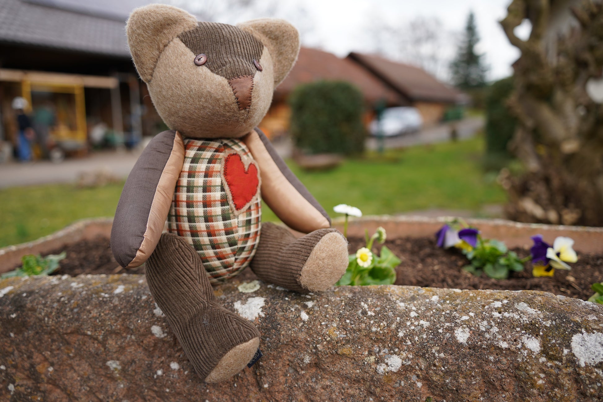 Ein brauner Teddybär aus verschiednenen Stoffen sitzt auf einem Blumenkübel mit bunten Veilchen im Hintergrund. Der Bär hat einen karierten Körper und ein rotes Herz auf der Brust.  