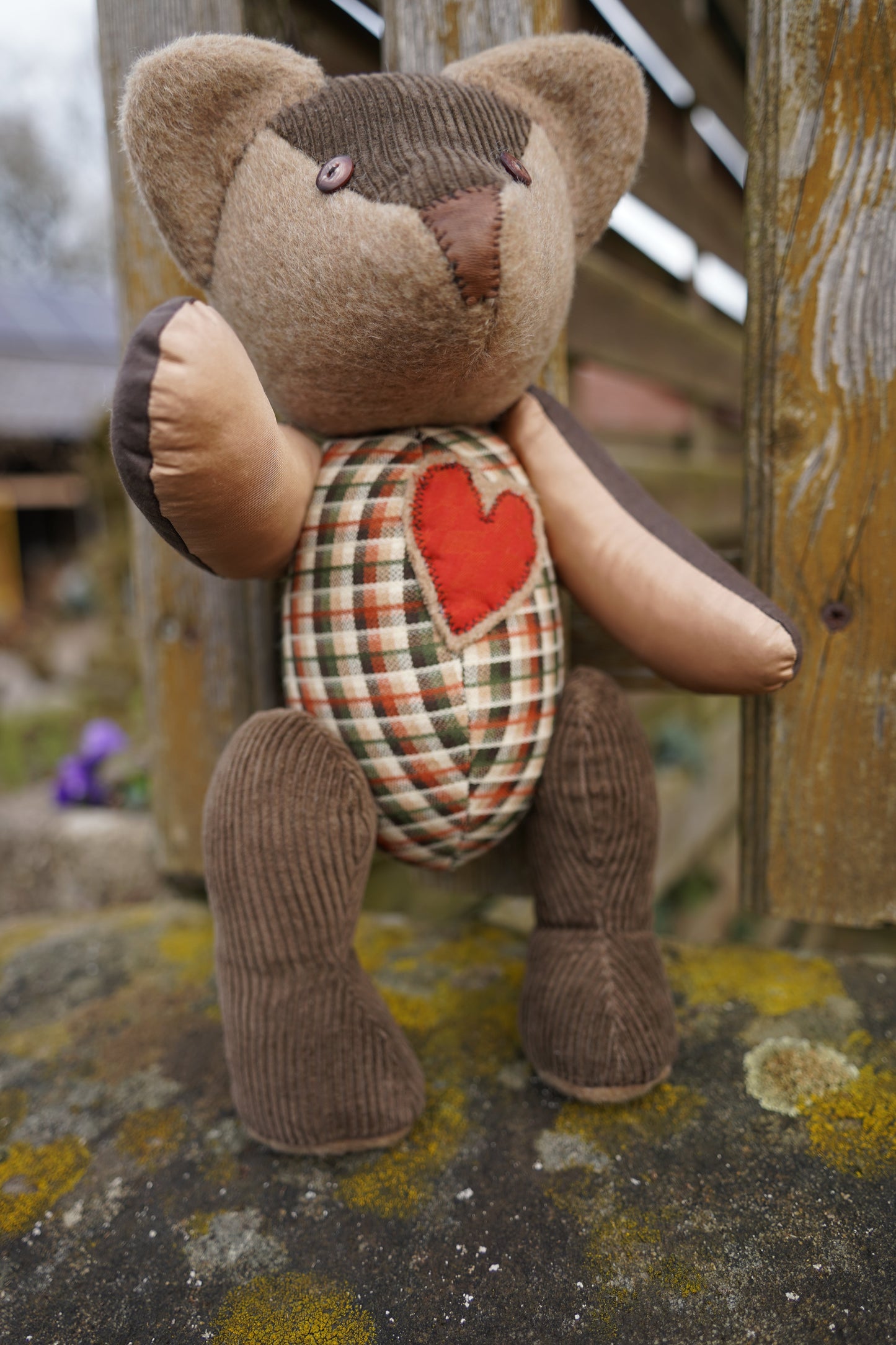 Ein brauner Teddybär aus verschiednenen Stoffensteht auf einer Mauer. Er hat einen karierten Körper und ein rotes Herz auf der Brust.  Der Bär sitzt auf einer Steinmauer. Er hat braune Knopfaugen und eine braune Ledernase. 