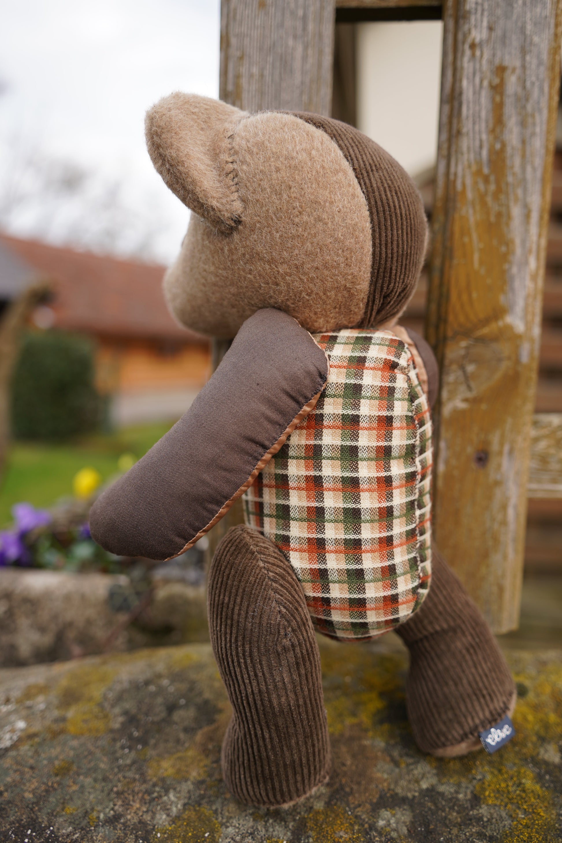 Ein brauner Teddybär aus verschiednenen Stoffen steht auf einer Mauer. Man sieht ihn von hinten.