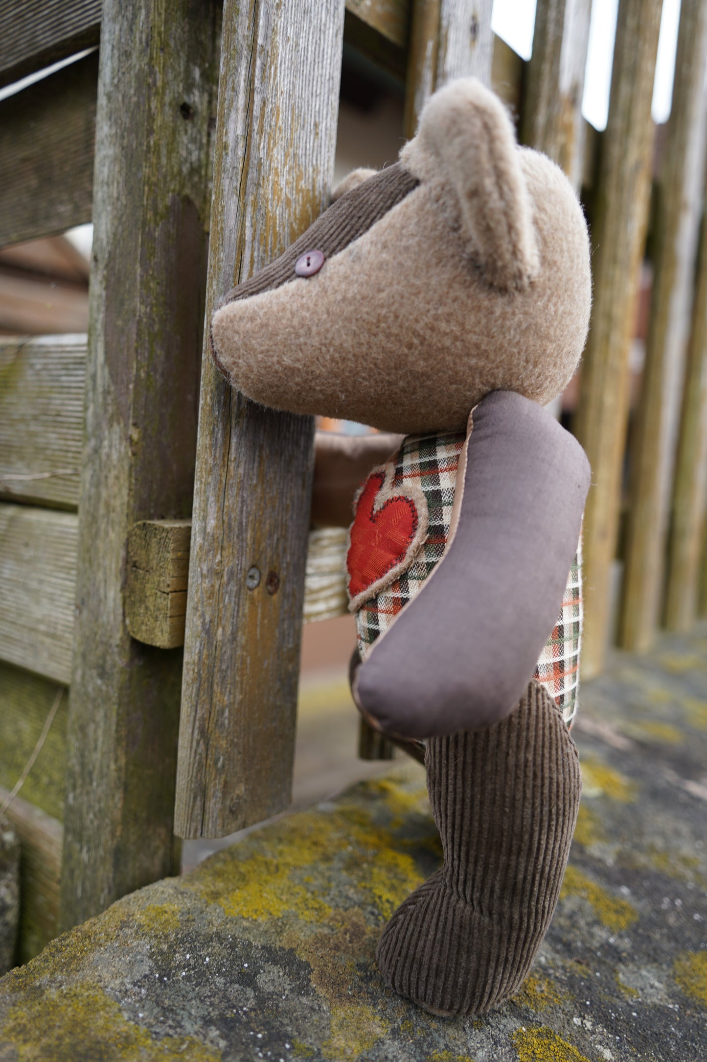 Ein brauner Teddybär aus verschiednenen Stoffen lehnt seitlich an einem Zaun Er hat einen karierten Körper und ein rotes Herz auf der Brust.  Er hat braune Knopfaugen und eine braune Ledernase. 