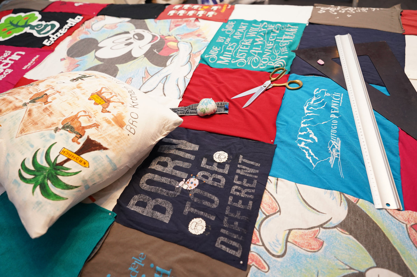 Eine Patchwork-Decke aus alten T-Shirts mit verschiedenen Designs und Texten. Darauf liegt ein Kissen aus einem T-Shirt, eine Schneiderschere, ein Nadelkissen mit Stecknadeln und ein Lineal.