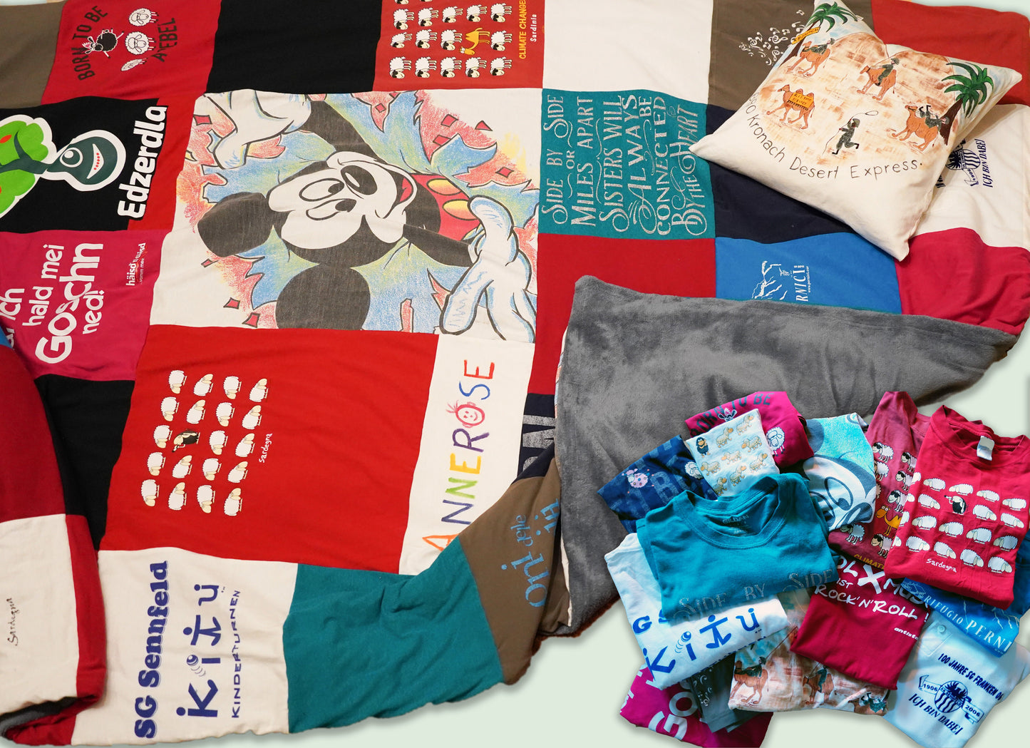 Eine bunte Patchwork-Decke aus verschiedenen T-Shirts, z.B. mit einem Mickey-Mouse-Design. Die Decke ist von einem Stapel gefalteter T-Shirts umgeben. Auf der Decke liegt ein Kissen, das aus einem T-Shirt genäht wurde.